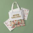 A white tote bag with "Merry Christmas" in gold letters and colorful gift illustrations at the bottom, placed on a green surface with sheets of matching present-themed stickers.