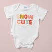 A white baby onesie on a light pink background with the phrase "SNOW CUTE" printed in colorful, patterned letters on the front.