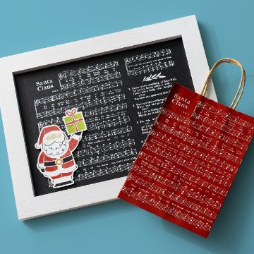 Show details for Christmas Music Sheet A framed sheet of Christmas music titled "Santa Claus" sits next to a red gift bag with the same sheet music design. A cute cutout of Santa holding a gift is attached to the frame, all on a blue background.