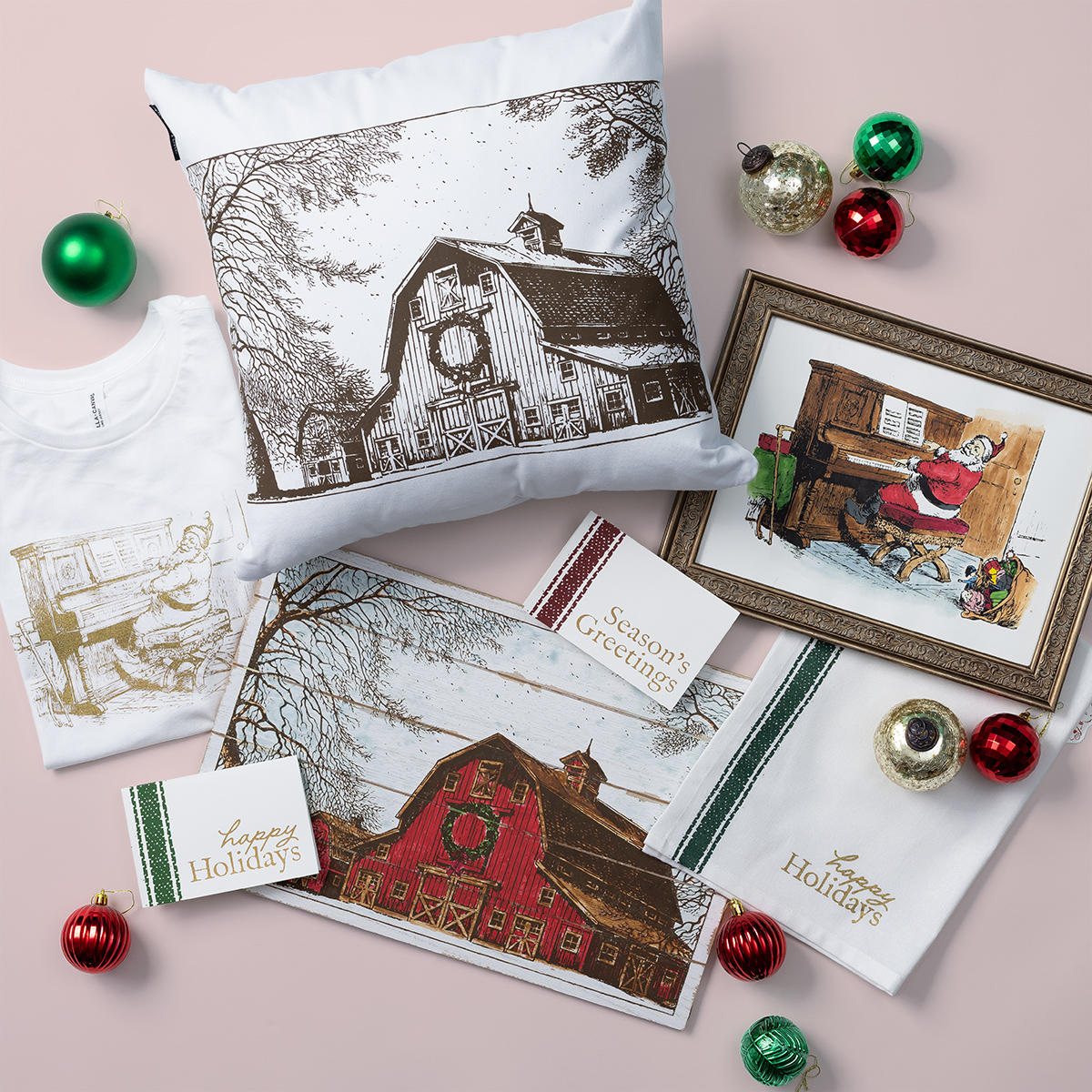 Holiday-themed items laid out on a pink surface, including a pillow, greeting cards, tea towels, a T-shirt, placemat, framed artwork, and Christmas ornaments, all featuring vintage barn and piano illustrations.