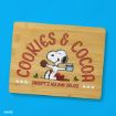 A wooden cutting board features Snoopy holding a cookie and a mug, with text reading “COOKIES & COCOA” and “Snoopy’s Holiday Treats,” decorated with holly and stars on a bright blue background.