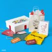 A white gift box with Peanuts characters, a white gift bag with Charlie Brown, gift tags, a yellow zigzag shirt, a greeting card with Snoopy, and tissue paper are arranged on a blue background.