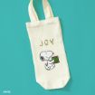 A cream-colored tote bag with "JOY" written above an illustration of Snoopy holding a green and brown book, set against a teal background.
