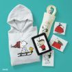 A gray hoodie with Snoopy and Woodstock on a sled, a tote bag with "JOY" and Snoopy, an e-reader with Charlie Brown, and various Snoopy-themed holiday cards on a teal background.