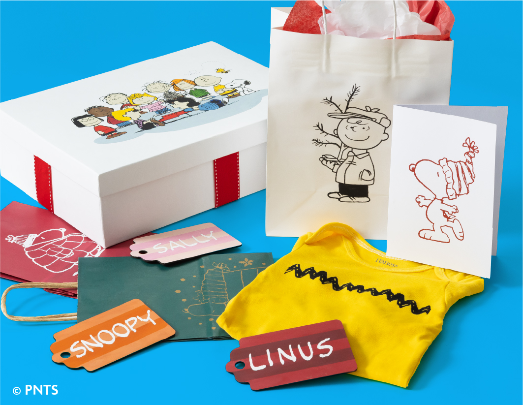 A Peanuts-themed gift set with a white box featuring Peanuts characters, a white gift bag with Charlie Brown, a card with Snoopy, a yellow Charlie Brown T-shirt, and name tags reading "Snoopy," "Saucy," and "Linus.