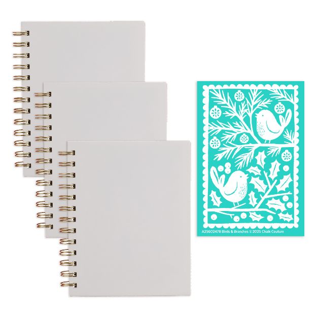 Three blank white spiral-bound notebooks are arranged in a stack next to a teal stencil featuring two birds, tree branches, leaves, and berries in a decorative folk art style.