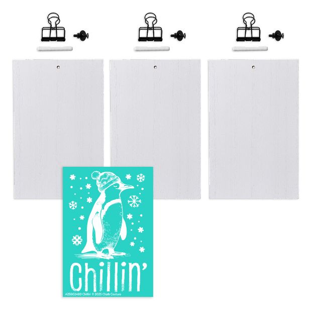 Three blank white wooden boards are arranged side by side with metal clips and mounting hardware above each. Below them is a teal card with a penguin wearing a beanie, snowflakes, and the word “Chillin’.”.