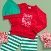 A festive children's outfit with a red sweatshirt reading "Santa's little helper," green and white striped pants, a green hat with a red brim, and Christmas-themed gift boxes on a green background.