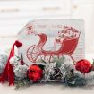 A holiday decoration shows a vintage postcard design with a red sleigh full of gifts, surrounded by frosted greenery, pinecones, red berries, and shiny red ornaments with a red ribbon.