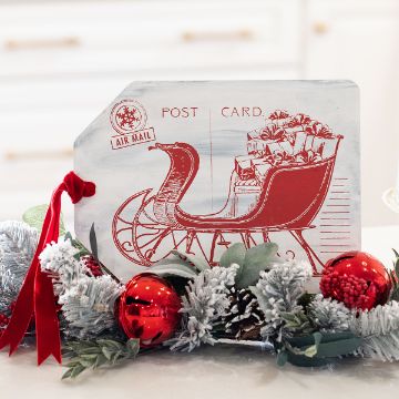 Show details for Debbie - Holiday Postcard A holiday decoration shows a vintage postcard design with a red sleigh full of gifts, surrounded by frosted greenery, pinecones, red berries, and shiny red ornaments with a red ribbon.