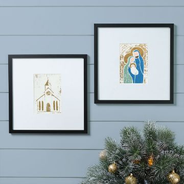 Show details for Debbie - Painterly Reverence Two framed art prints hang on a light blue wall; one depicts a church, the other shows a stylized Nativity scene. Below, a Christmas tree with gold ornaments is partially visible in the corner.