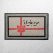 A rectangular doormat with a red ribbon and bow design, featuring the word "Welcome" in cursive script on a beige background with a black border.