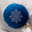 A round blue pillow with a white snowflake design in the center, placed on a light surface near a cozy blanket and a gift wrapped with a gold ribbon and handwritten note.