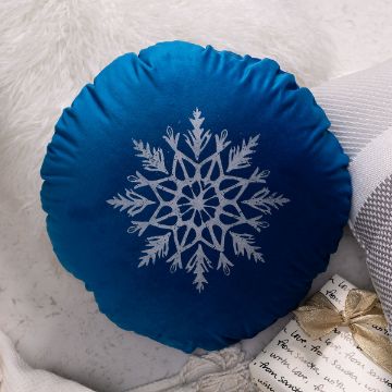 Show details for Debbie - Farmhouse Snowflake A round blue pillow with a white snowflake design in the center, placed on a light surface near a cozy blanket and a gift wrapped with a gold ribbon and handwritten note.