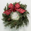 A green pine wreath with pinecones and a large red plaid bow hangs on a white wall. In the center is a sign that reads "Peace on Earth" with decorative foliage and berries around the text.