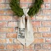 A green wreath hangs on a brick wall with a beige cloth bow that has "O Holy Night" written in decorative script and a star design printed on the hanging tails.
