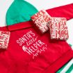 A red and green hoodie with "Santa's little helper" printed on it lies on a white surface, surrounded by three small, gift-wrapped presents with festive red and white holiday designs.