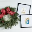 A green holiday wreath with pinecones, red plaid bow, and “Peace on Earth” sign is next to two framed art prints: one of a church, and one of Mary, Joseph, and baby Jesus. All are displayed on a white wood wall.