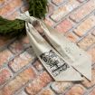 A beige cloth ribbon with “O Holy Night” and star designs is tied in a knot and draped over green fir branches, resting on a pink and tan brick surface.