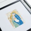 A framed artwork depicts a stylized Holy Family with gold halos: Mary in green, Joseph in blue, and baby Jesus in white, set against a white background.