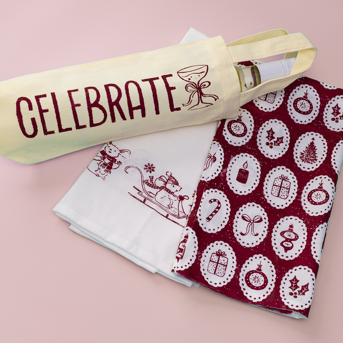 A wine bottle in a yellow bag labeled "CELEBRATE" sits next to two festive tea towels with holiday-themed prints, one white with red designs and one maroon with white Christmas icons, on a pink background.