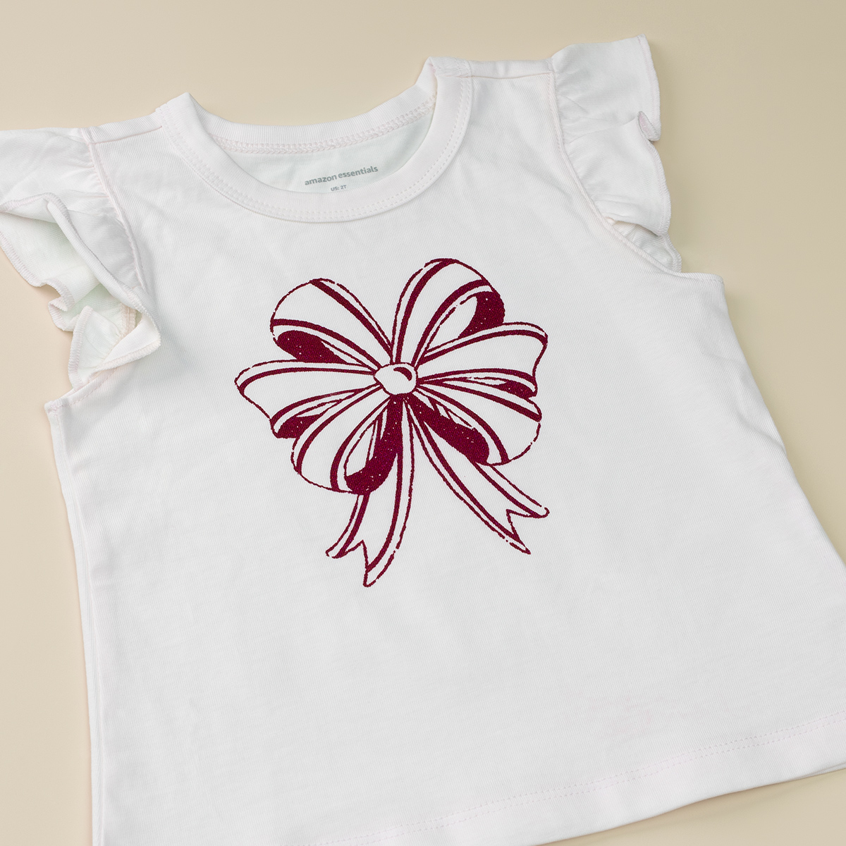 White short-sleeve children’s shirt with ruffled sleeves, featuring a large maroon bow illustration on the front, laid flat on a light beige background.