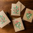 Four brown holiday cards with scalloped edges are decorated with holly and mistletoe illustrations. A gold pen and an open card box are placed nearby on a wooden surface.
