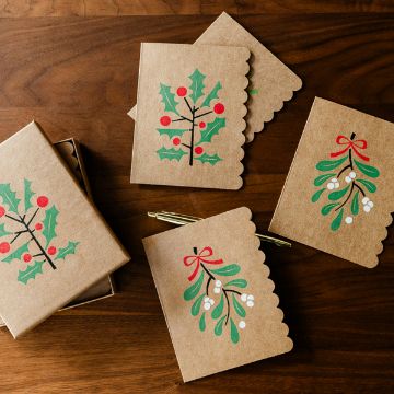Show details for Under the Mistletoe Four brown holiday cards with scalloped edges are decorated with holly and mistletoe illustrations. A gold pen and an open card box are placed nearby on a wooden surface.