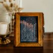 A wooden-framed picture with a bark edge displays white line art of tall pine trees and stars against a dark blue night sky, set on a wooden table beside a glass candle holder with a white candle.