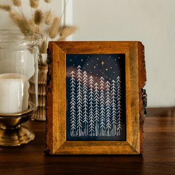 Show details for Enchanted Forest A wooden-framed picture with a bark edge displays white line art of tall pine trees and stars against a dark blue night sky, set on a wooden table beside a glass candle holder with a white candle.
