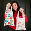 A woman in a red sweater smiles and holds up two festive gift bags. One bag features colorful ornaments, and the other has the words "OH WHAT FUN" in red, green, and gold letters.