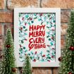 A framed holiday print with colorful Christmas lights and the words "Happy Merry Everything" in red, surrounded by small decorated Christmas trees, set against a brick wall background.