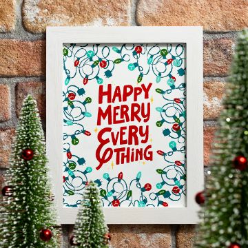 Show details for Happy Merry Everything A framed holiday print with colorful Christmas lights and the words "Happy Merry Everything" in red, surrounded by small decorated Christmas trees, set against a brick wall background.