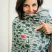 A woman smiles while pointing at a gray scarf with green tree and forest designs, featuring a small red pattern among the prints.
