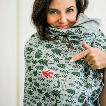 Show details for Wild Things A woman smiles while pointing at a gray scarf with green tree and forest designs, featuring a small red pattern among the prints.