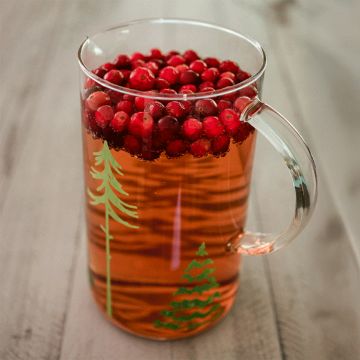 Show details for Pining For You A clear glass mug with green tree designs is filled with a reddish drink, topped with fresh cranberries, and placed on a light wooden surface.