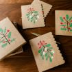Brown holiday cards with scalloped edges and festive leaf illustrations, including holly and mistletoe, are spread out on a wooden table, along with a gold pen and a matching card box.