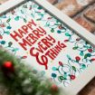 A framed festive print with the words "Happy Merry Everything" in red surrounded by colorful holiday lights, placed on a brick surface with blurred greenery and a red ornament in the foreground.