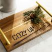 A wooden tray with gold handles displays the words "COZY UP," a drawn mug, and a small pine branch with pinecones, placed on a light surface next to a white mug.