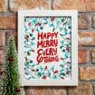 A white-framed print with colorful holiday lights and the words "HAPPY MERRY EVERY THING" in bold red letters, propped against a brick wall near a small decorated Christmas tree.