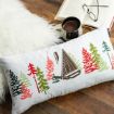 A rectangular pillow with colorful trees and a cabin design rests on a wooden floor beside a fluffy white rug, an open book, and a mug of hot chocolate.