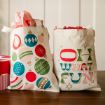 Two festive tote bags sit on a wooden surface. One features colorful Christmas ornaments, while the other has bold, retro-style text reading “OH WHAT FUN” with stars. Both bags are filled with tissue paper.