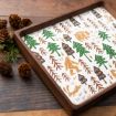 A wooden tray with a white base featuring illustrations of green trees, a brown bear, a Bigfoot figure, and an otter. Pinecones and greenery are arranged beside the tray on a wooden surface.