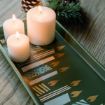 Three lit white candles sit on a green tray with gold and white geometric patterns. In the background, there is a pine cone and some pine branches on a wooden surface.