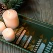 Three lit white candles sit on a green tray decorated with images of stylized candles. A pinecone and pine sprig rest nearby on a wooden surface, creating a cozy and festive atmosphere.