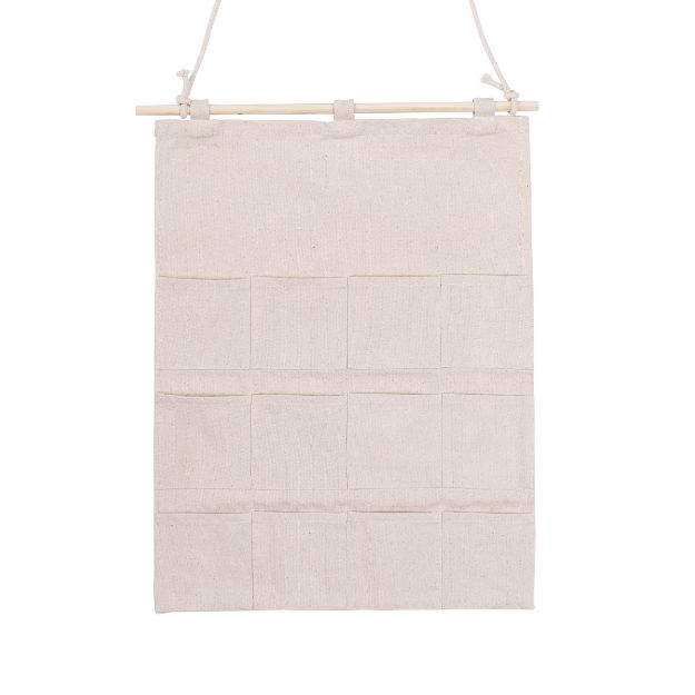 A beige fabric wall organizer with twelve small pockets arranged in three rows, hanging from a wooden dowel by a string.