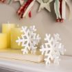 White decorative snowflakes rest on a closed book beside three unlit yellow candles. In the background, a garland of patterned stars and ribbons hangs along the wall, adding a festive touch.