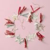 A star-shaped garland made of patterned paper stars and assorted red, green, and white ribbons is arranged in a circle on a pink background, with a piece of twine attached for hanging.