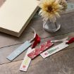 Three festive bookmarks with Christmas designs and red or white ribbons lie on a wooden surface near a closed book and a small glass vase holding cream-colored flowers.