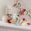 A festive mantel displays a gift bag with “He sees you when you’re drinking,” decorative snowflakes, a gold ornament, a red ornament, a stack of books, holiday bookmarks, and a string of star-shaped garland.